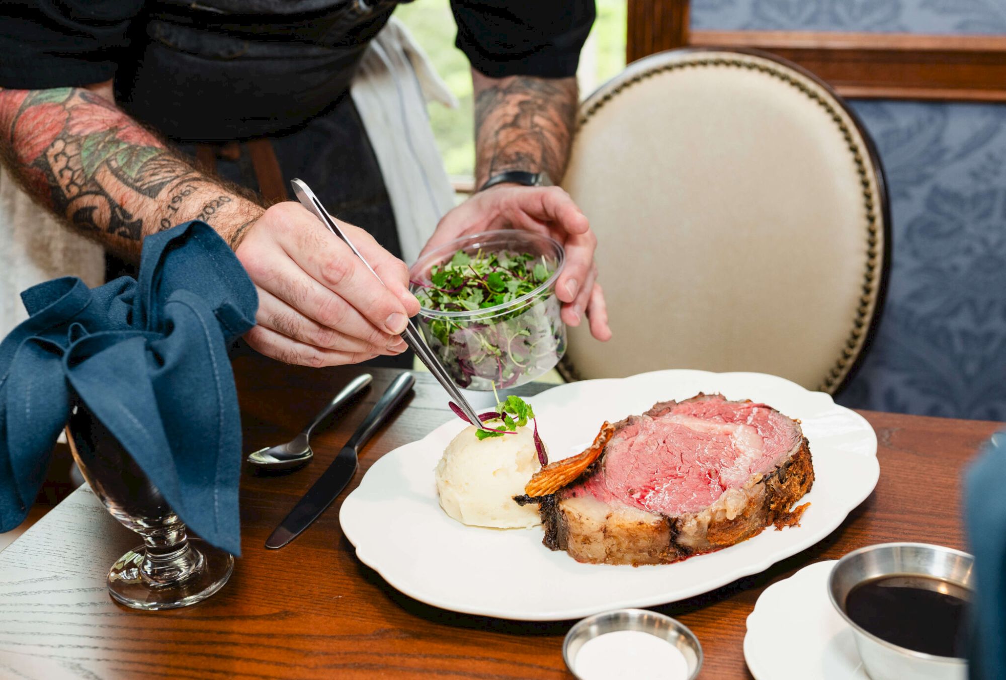 A chef or diner adds green herbs onto a slice of pink roast beef on a white plate, with bread and a glass of red wine nearby, at a dining table.