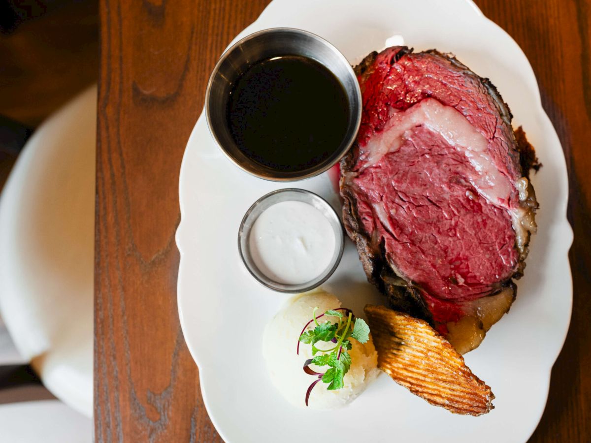A plated herb-crusted roast beef steak with red center, dark jus, a small ramekin of sauce, a dollop of white crema, a potato chip, and greens on a white plate, photographed from above.