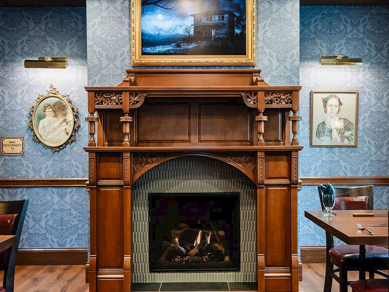 A traditional wood fireplace mantel with ornate carvings, a framed painting above, vintage circular clock and portrait on blue patterned walls, and wooden tables and chairs in a cozy dining room.