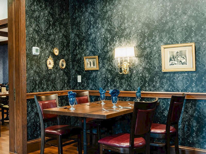 A cozy dining nook with a dark teal patterned wallpaper, wooden table, red-cushioned chairs, wall art, and warm lighting.