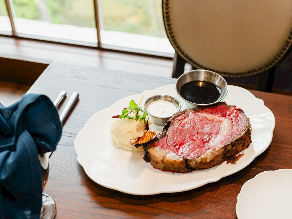 A plated steak with pink center, a small potato croquette, greens, and sauces on a white plate at a table by a window.