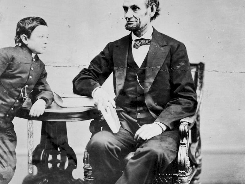 A man in a suit sits in a chair, reading a newspaper, beside a child at a small table, both looking toward each other.