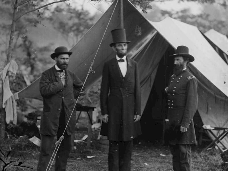 Three men in 19th-century attire stand in front of a tent camp, likely Civil War era, posing for a photo with a flagpole nearby.
