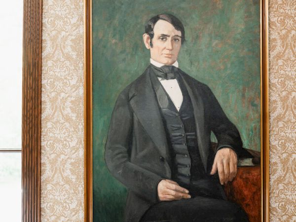 A Victorian-era man portrait in a dark suit, seated, with a calm expression, framed in a gold frame against a patterned wall.