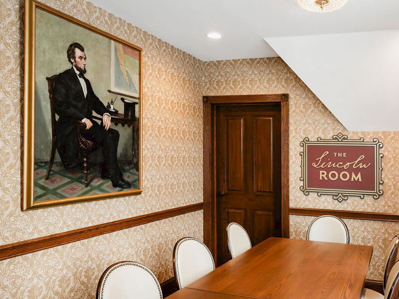 A formal meeting room with a long wooden table, white chairs, ornate wallpaper, a large portrait on the wall, and a sign reading &ldquo;The Lincoln Room&rdquo; beside a wooden door.