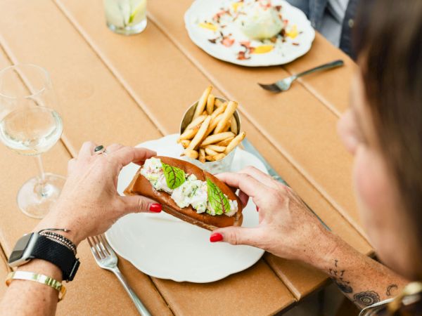 Two people at a table: a person ahead placing lettuce-topped burger together, french fries on plate, wine glasses and salad in the background, casual lunch.