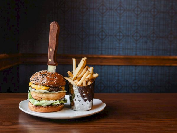 A burger with lettuce and cheese on a plate, a side of French fries in a white cup, and a large steak knife stabbed into the sandwich on a wooden table.