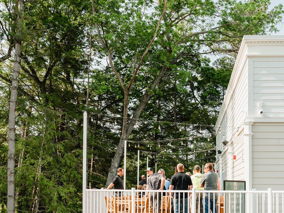 A group of people gather on a white deck or balcony near a modern white building, surrounded by tall green trees on a sunny day.