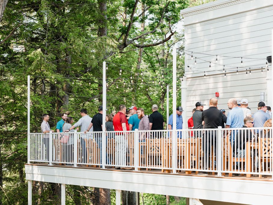People on a wooden balcony/deck attached to a white house, chatting in a line among trees, outdoor gathering with string lights.