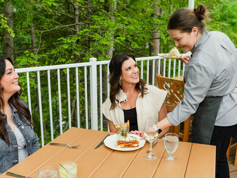 Three friends dine outdoors as a waitress serves food and drinks at a wooden table on a sunlit patio.