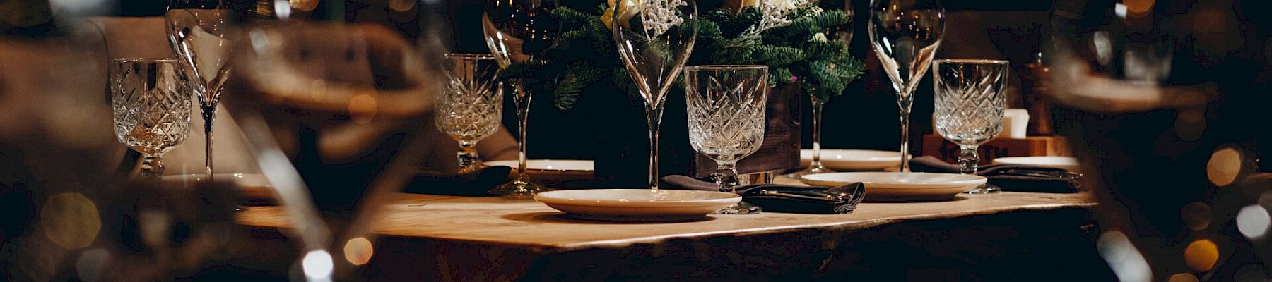 A dimly lit dining table set for a formal meal with champagne flutes, crystal glasses, plates, and greenery centerpiece, festive and elegant.