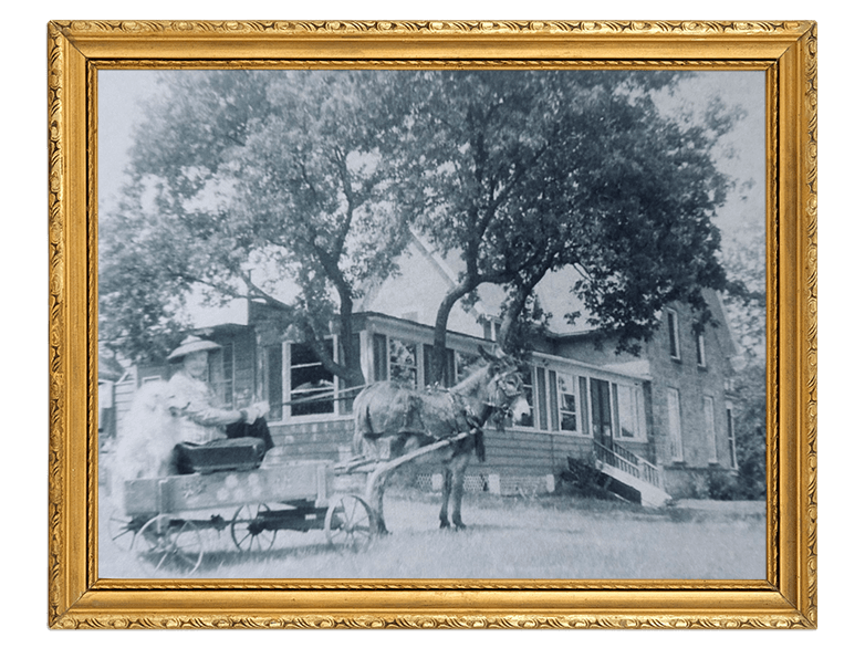 A vintage black-and-white photo framed in gold shows a horse-drawn carriage in front of a house with trees; a person stands near the carriage.
