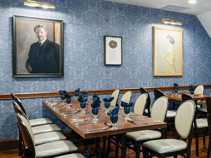 A formal dining room with a long wooden table set for guests, blue patterned wallpaper, framed portraits, and elegant cream cushions on dark wood chairs.
