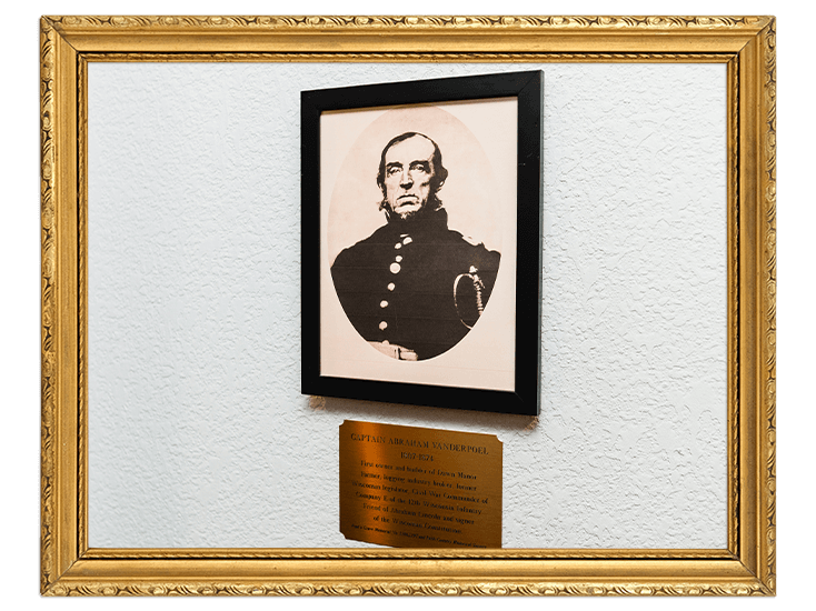 Old black-and-white portrait of a stern man in a dark military coat, framed, with a plaque underneath in a gold ornate frame.