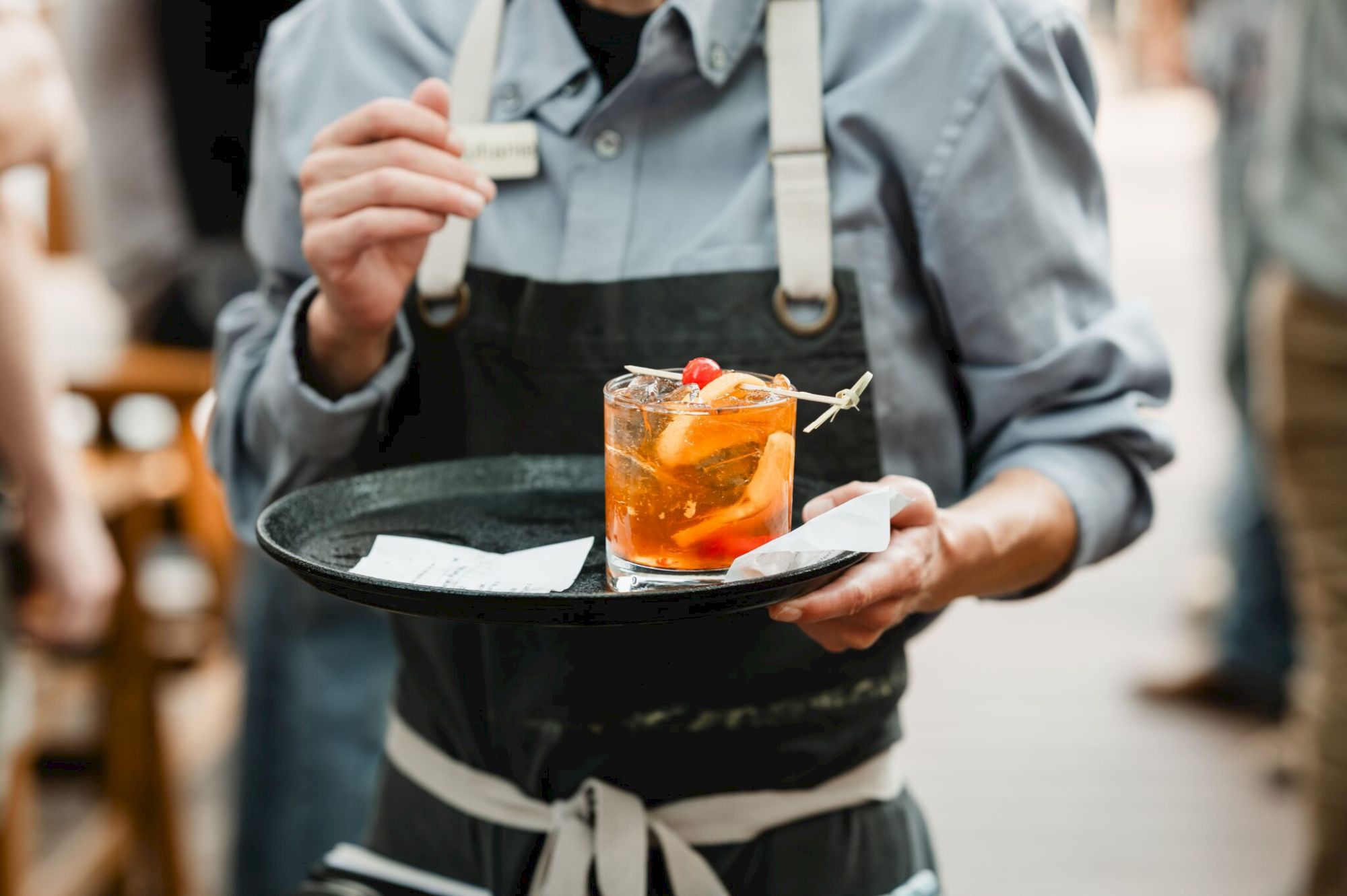 A waiter in a gray shirt and black apron holds a tray with a drink garnish and a small napkin, presenting it to a guest.