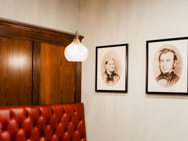 A cozy corner with a tufted red leather booth, warm wooden wall paneling, a hanging lamp, and two vintage black-and-white framed portraits on the wall.