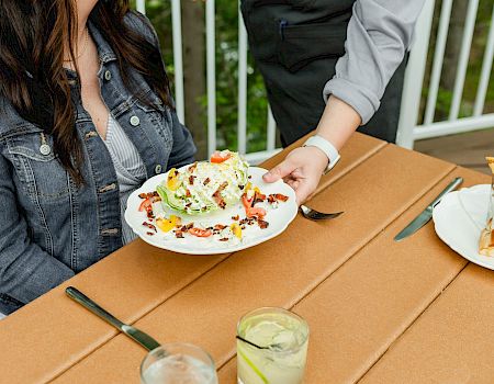 A couple enjoys a stylish outdoor meal: plates of colorful salads and sandwiches, plus drinks on a sunny wooden table, shared chatter. g