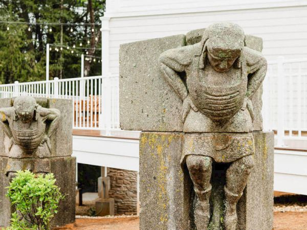 Stone carved statues of crouching humanoid figures beside a white building; one larger in front, another smaller behind, outdoors.
