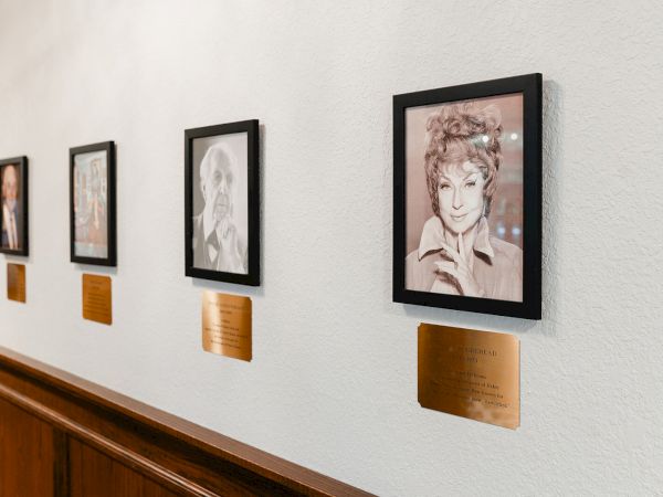 A gallery wall with framed black-and-white portraits along a light textured wall, each with a small bronze plaque beneath.