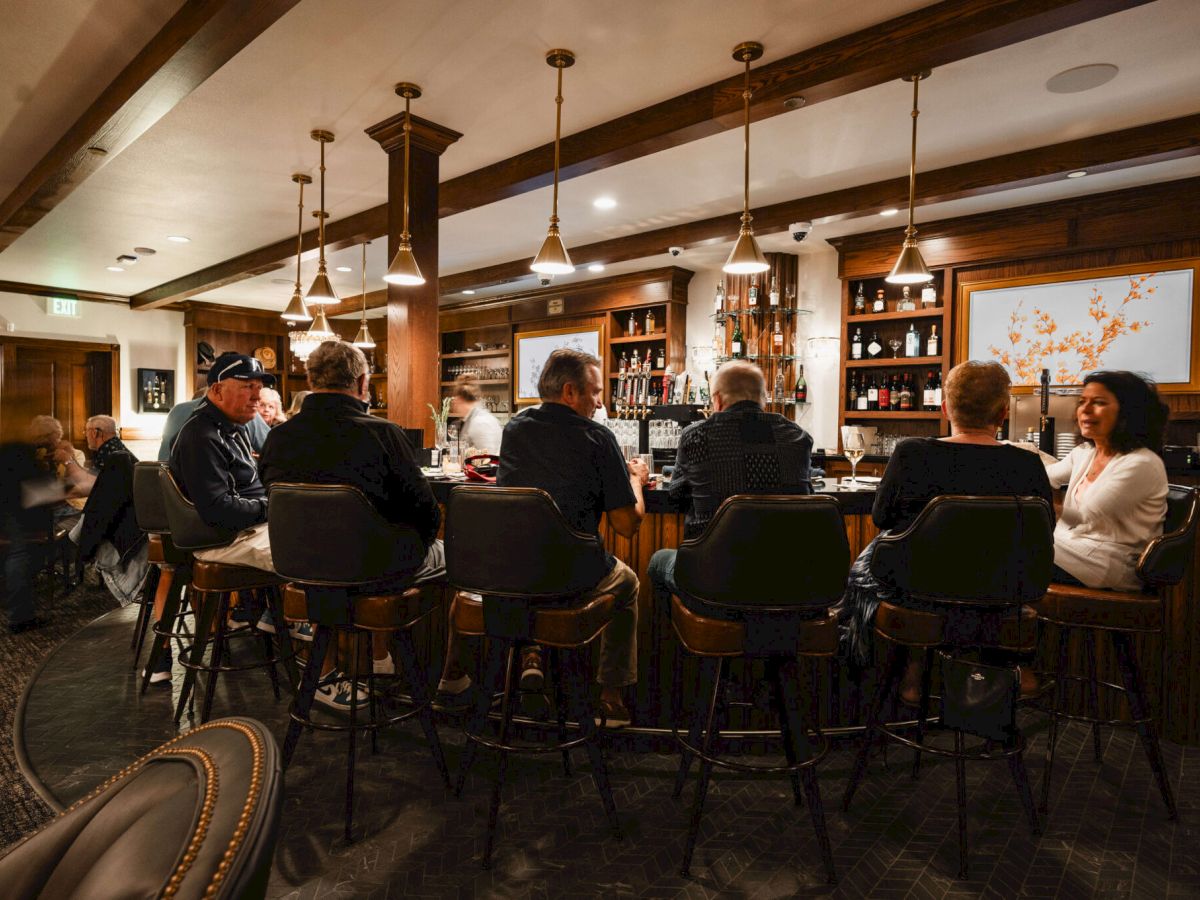 A cozy upscale bar or lounge with a long wooden counter, patrons seated on high chairs, warm lighting, and shelves of bottles behind the bar.