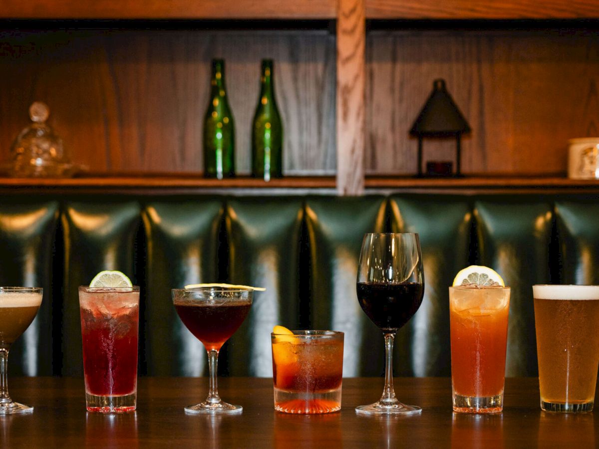 A row of seven assorted cocktails on a bar counter, with bottles and decor on the shelves behind, ending with a dark leather booth.
