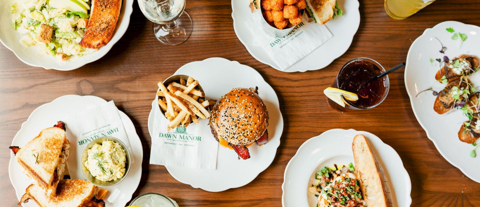 A variety of plates with burgers, fries, salads, soups, and drinks spread across a wooden table, resembling a casual restaurant feast.
