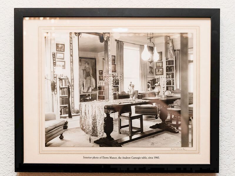 A framed black-and-white photo of a cozy, sunlit room with a table, chairs, and shelves, mounted on a wall.