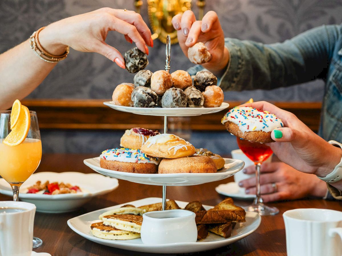 A lavish tea-time spread: scones, pastries, donuts, and drinks, with hands reaching to grab treats.