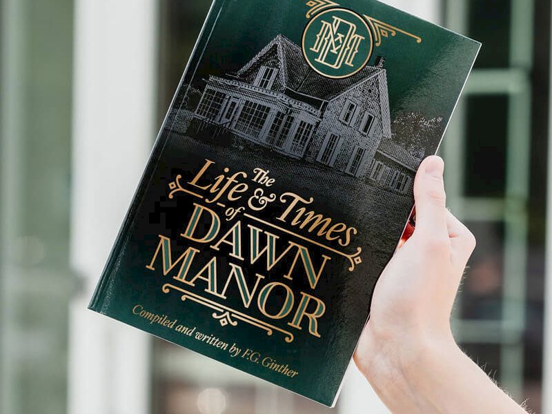 A person is holding a dark green book titled &ldquo;The Life & Times of Dawn Manor&rdquo; with gold lettering and a building illustration on the cover, indoors.