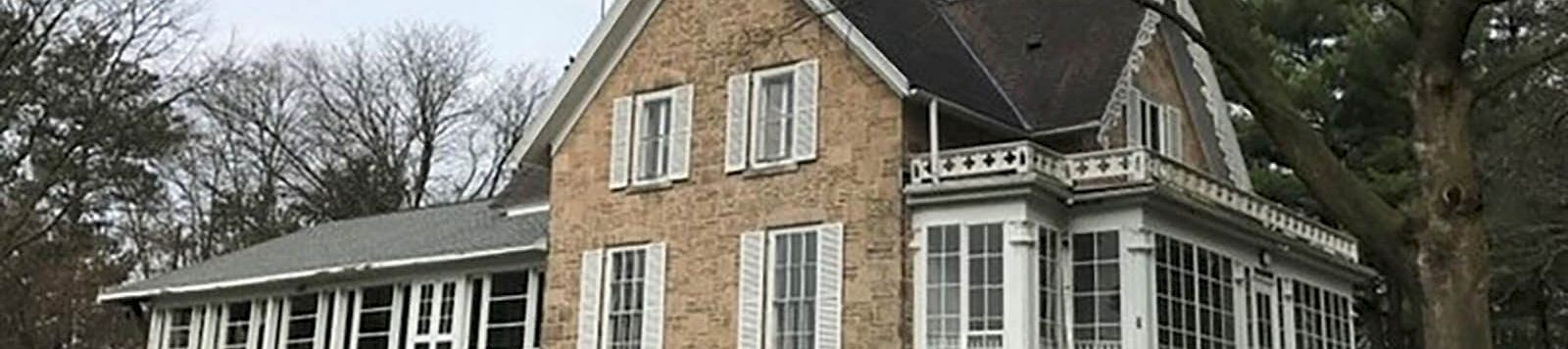 A large stone house with white-trimmed windows, a sunroom, and an attached glass-enclosed porch, set on a grassy lawn under bare trees.
