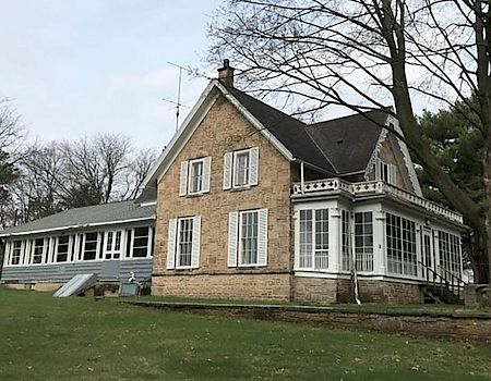 A brick house with a glass-enclosed porch, surrounded by trees on a grassy lawn, under a cloudy sky.