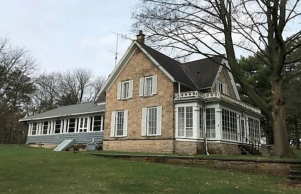 A brick house with a glass-enclosed porch, surrounded by trees on a grassy lawn, under a cloudy sky.