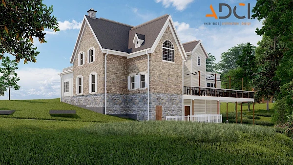 A two-story stone house with a steep gabled roof sits on a grassy hillside, featuring a wraparound deck and large windows under a sunny sky.