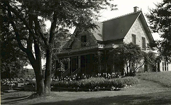 Old white house with a wrap-around porch, a garden in front, and people gathered on the lawn near the entrance.