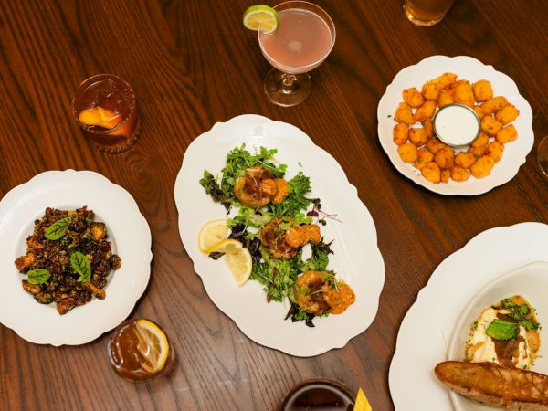 A top-down spread of assorted dishes: fried bites, greens with citrus, crusty bread, and small plates with dipping sauces, plus drinks on a wooden table.
