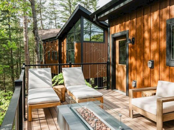 A wooden deck with lounge chairs and a glass coffee table extends from a cabin, overlooking a lush green forest.