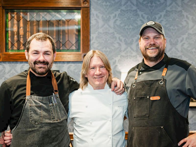 Three smiling chefs stand together in a kitchen, wearing aprons; a woman in the middle wears a white chef coat.