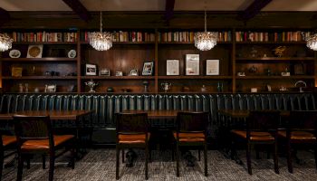 A cozy, dark library bar with a long wooden shelf wall, upholstered chairs, and hanging chandeliers, creating a warm, intimate reading space.