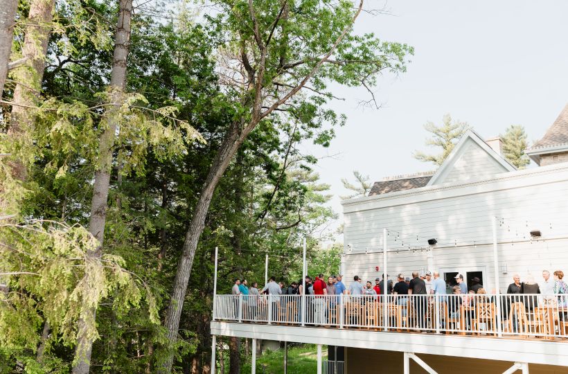 A crowd of people on a wooden balcony overlooking a green, tree-filled yard beside a light-colored building.
