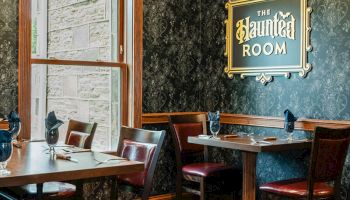 A cozy dining area with dark wood furniture, a chalkboard-style wall, and a framed sign reading &ldquo;The Inn Room,&rdquo; near a window.