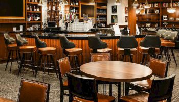 A cozy bar with wooden round tables and cushioned chairs, a long counter lined with stools, warm lighting, and shelves of bottles in the background.