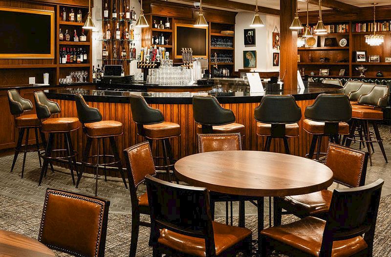 A cozy bar with wooden round tables and cushioned chairs, a long counter lined with stools, warm lighting, and shelves of bottles in the background.