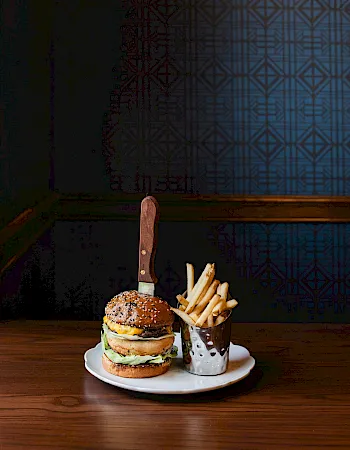 A plate with a tall burger, fries in a mini cup, and a long breadstick upright; dark patterned wall behind on a wooden table.