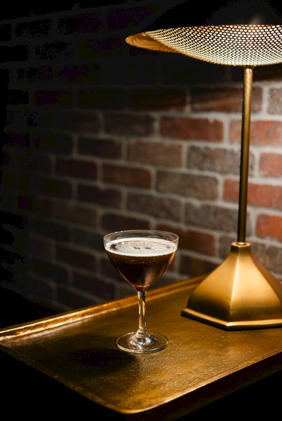 A cocktail in a coupe glass beside a modern gold lamp on a wooden surface, with a brick wall backdrop.