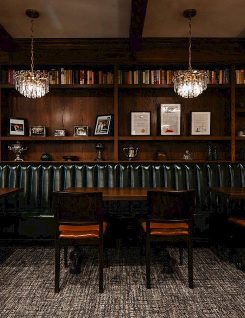 A dark, wood-paneled library bar with high-backed chairs facing a long counter, shelves full of books, framed photos, and warm pendant lights.