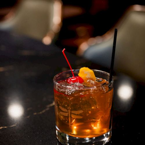 A short, 140-character description: A glass tumbler with amber cocktail, ice, a bright red cherry, and a lemon twist garnish, on a dark bar counter.