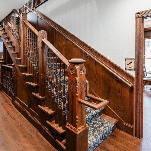 A wooden staircase with dark railings, carved balusters, and a turned newel post, leading up beside a carpeted runner.