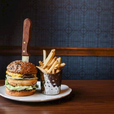 A cheeseburger with a big knife stuck in it and a side of French fries in a metal cup on a white plate, all on a wooden table.