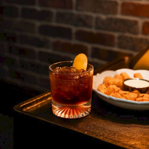A slice of toast with butter and jam, a glass of red juice, and a plate on a brick counter with dim lighting.