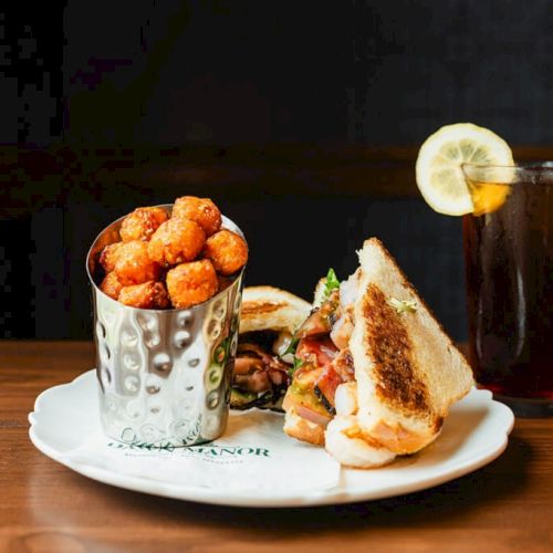 A plate with a metal cup of fried cheese balls, a toasted sandwich, and a glass of iced tea with lemon on a wooden table.